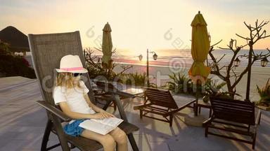 年轻的白种人女孩坐在扶手椅上看书。 海上日落。 长椅子和雨伞。 有<strong>相同</strong>的剪辑