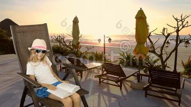 年轻的白种人女孩坐在扶手椅上看书。 海上日落。 长椅子和雨伞。 有<strong>相同</strong>的剪辑