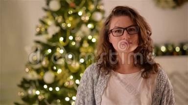 美丽的女孩，长着卷发和眼镜，看着相机和微笑的圣诞树背景。 特写