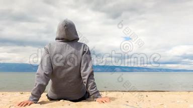 电影摄影-男人或女人坐在海滩上的帽衫里。 后景。 4k