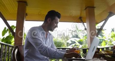 在热带森林的夏季<strong>露台</strong>酒店，年轻的商人在早餐时用笔记本电脑工作，小伙子一边喝果汁