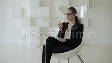 商务女士坐在一张白色椅子上，上面放着一台电脑平板电脑，在办公室里用手机说话