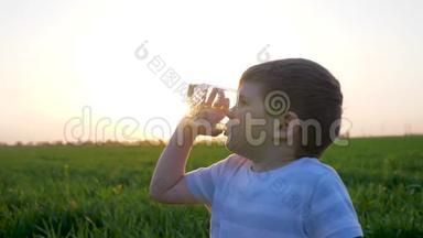 草地上的小男孩手里拿着玻璃杯，喝着矿泉水，孩子坐在绿茵场上，孩子喝着水
