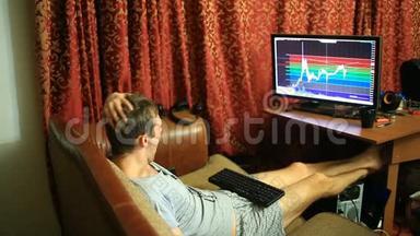 一个穿着T恤和短裤的男人，坐在沙发上的家里，监视着货币兑换时间表的变化
