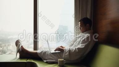年轻的微笑商人在手提电脑上工作，穿着<strong>白色浴袍</strong>，坐在窗边喝杯咖啡
