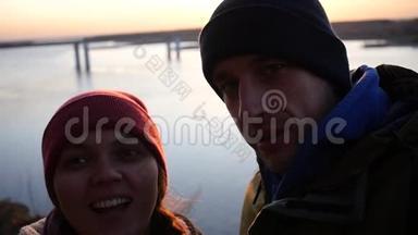 年轻夫妇正在电话里拍照，在<strong>河边</strong>的夕阳<strong>背景</strong>下笑着看着摄像机