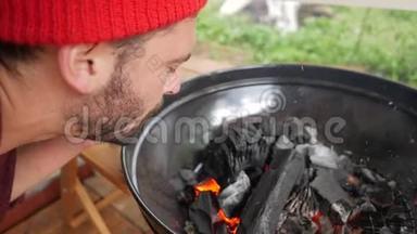 年轻的大胡子在烧烤炉里吹着煤火. 黑色木制木炭燃烧在Bbq。 高清高清