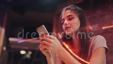 年轻迷人的女人，美丽的微笑是使用智能手机在晚上。 坐在<strong>酒吧</strong>或<strong>餐馆</strong>里的女孩