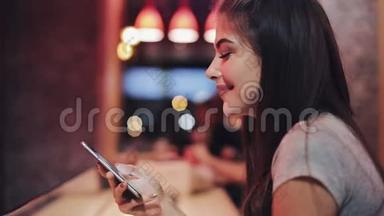 年轻迷人的女人，美丽的微笑是使用智能手机在晚上。 <strong>坐在</strong>酒吧或<strong>餐馆</strong>里的女孩