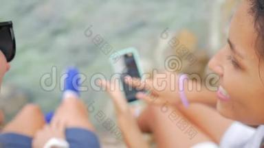 两个无忧无虑的混合赛跑模特坐在海边的悬崖岩石上，在手机上观看照片。 生活水平高清