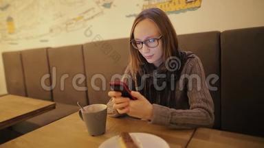 戴眼镜的女人在咖啡馆里使用智能手<strong>机上</strong>的应用程序，喝咖啡，微笑着在手<strong>机上</strong>发短信。 年轻漂亮