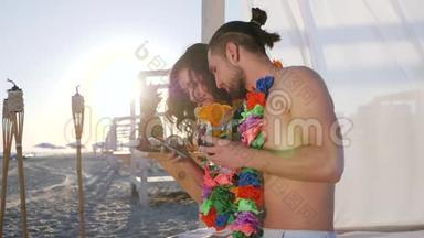 来自夏威夷的年轻人脖子上挂着安卓，女孩和男孩坐在沙滩上，手里拿着手机
