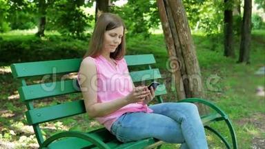 年轻女子坐在公园的长凳上，用智能手机。 在露天使用智能手机的漂亮女孩