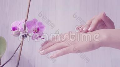 嫩女人手的特写镜头涂抹护肤霜.. 紫色兰花的光背景。 美丽的双手优雅