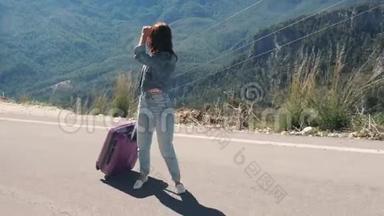 美丽的年轻女士在农村的道路上，带着手提箱搭便车在阳光明媚的日子户外