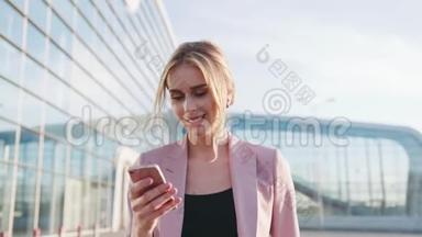 一个自信的年轻金发女孩穿着一件优雅的粉红色夹克，走在机场航站楼旁，用她的手机