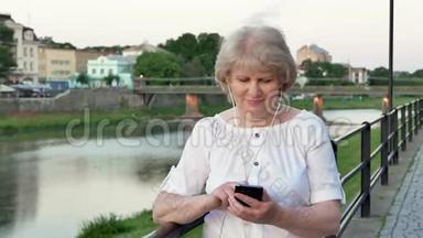 傍晚时分，在海滨、老城区、河<strong>边</strong>和<strong>桥边</strong>，使用带有耳机的智能手机的老年妇女