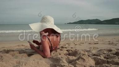 年轻女子在海滩上晒日光浴时用手机。 穿着比基尼和太阳帽的漂亮女孩拍照