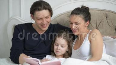 年轻的一家人正在读一本小女孩躺在床上的书。 母亲和父亲正在教他们的女儿