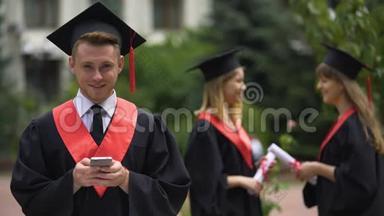 年轻的男毕业生拿着智能手机，微笑着拍照，毕业典礼