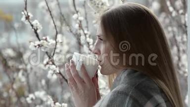 女孩站在窗前的阳台上，在盛开的杏树的背景下喝茶