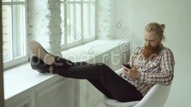 有<strong>胡子</strong>的年轻时髦<strong>男士</strong>在坐在椅子上时用智能手机发短信，双腿放在窗台上