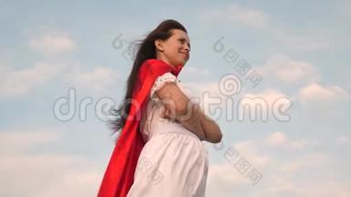 超级英雄女孩穿着红色斗篷站在田野上，斗篷在风中飘扬。 特写镜头。 女孩梦想成为一个