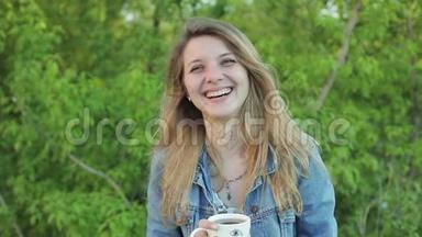 可爱的长发女孩笑着，手里拿着一个杯子在绿色灌木丛的背景下。 穿牛仔裤的漂亮金发女孩
