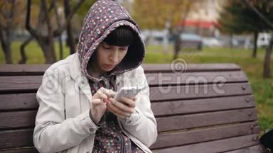 一位身穿连帽衫的年轻女子在秋<strong>季</strong>公园使用智能<strong>手机</strong>。