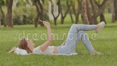 一个年轻的女孩躺在草地上，手里拿着一部智能手机。 在摄像机手机上摆姿势很有趣。 娱乐