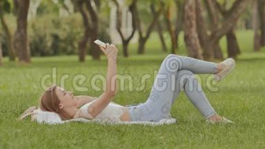 一个年轻的女孩躺在草地上，手里拿着一部智能手机。 在摄像机手机上摆姿势很有趣。 娱乐