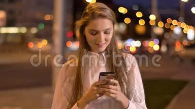 年轻有魅力的女孩正在夏天的市中心晚上在智能手机上输入信息，这是一个沟通概念