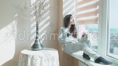 快乐的年轻女子坐在阳光下的窗台上。 一个美丽的女孩坐在窗前的阳光下