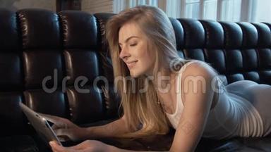 漂亮的年轻女人用应用平板电脑躺在皮革沙发上。 一个学生用<strong>手</strong>可爱地<strong>捂</strong>住她的<strong>嘴</strong>