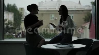 办公室窗口附近的两个商务妇女在一天结束时喝着咖啡，和热热闹闹的流言蜚语交谈