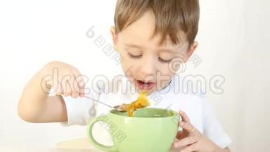 孩子高兴地用勺子从盘子里吃汤，<strong>特写</strong>。 一个男孩坐在白色背景的桌子旁<strong>吃饭</strong>