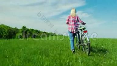 一个骑自行车的女人在风景如画的地方散步-一片绿色的草地和明亮的蓝天。 后景。 复制空间