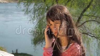 在水边拿电话的小女孩。 这孩子在露天。 一个女孩在阳光明媚的春天在河边。
