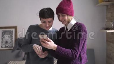 两个年轻的女人站在房间里看手机。 女人拿着智能手机