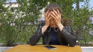 悲伤的漂亮女孩坐在咖啡馆的桌子旁。 在智能手机上读取短信。 双手抱着他的头悲伤