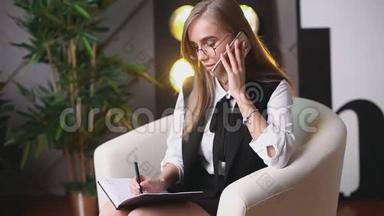 戴眼镜的年轻美女坐在办公室里打电话，在<strong>笔记本</strong>上说和写。 <strong>上面</strong>的植物