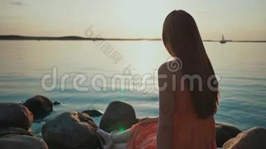 <strong>一个</strong>年轻的女孩坐<strong>在</strong>海滩上的岩石上。 周围没有人。 日<strong>落在</strong>潮湿的沙子和海水中反射。 关于