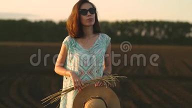 戴太阳镜的女孩站在日落时的田野里，拿着<strong>金色</strong>的<strong>麦穗</strong>和草帽。 乡村户外场景
