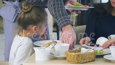 可爱的小女孩在意大利餐馆的意大利面条里倒奶酪，而她的年轻父母却吃他们的蔬菜