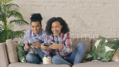 慢动作两个混合种族卷曲女孩朋友坐在沙发上玩控制台电脑游戏与游戏本和乐趣