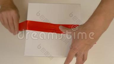 包装白色礼品盒。高加索人手拿包装礼盒。男人用手在一个白色的纸板箱周围系上一条红丝带