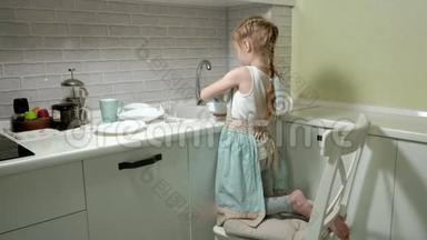 穿着围裙的漂亮小女孩站在明亮<strong>厨房</strong>的椅子上，用水<strong>冲洗</strong>盘子，帮助父母