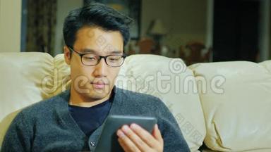 一个带眼镜的年轻正宗亚洲男人。 他坐在家里的沙发上看电子书