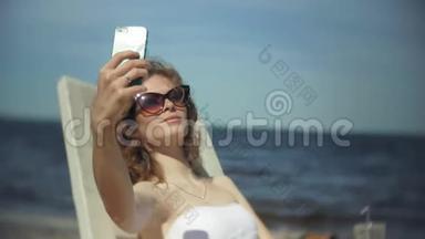 一个穿着白色比基尼的年轻女孩躺在<strong>海边</strong>沙滩上的休息室里<strong>晒太阳</strong>，并使用智能手机
