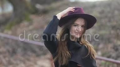 时尚开朗的年轻女孩留着长发，紫色的帽子和黑色的外套走在街上。 多云多<strong>风天</strong>气